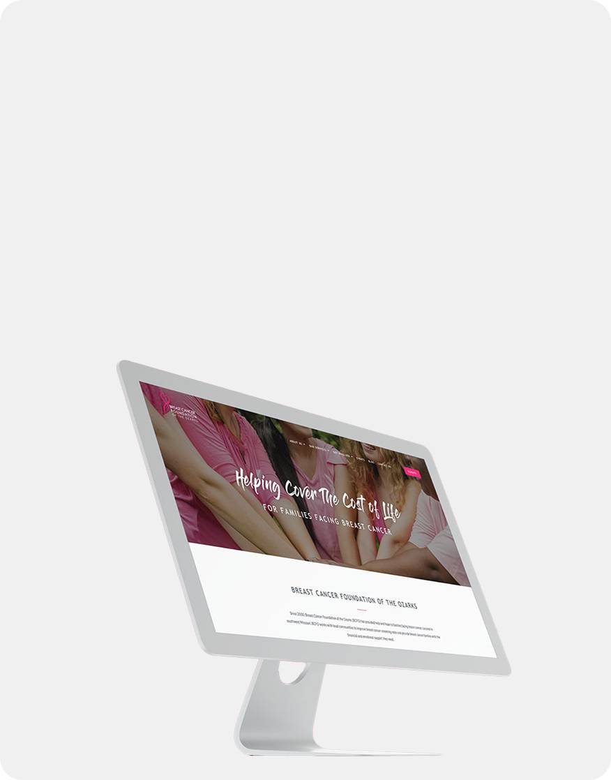 A desktop monitor displays a website with the text “Helping Cover the Cost of Life” and images of people holding hands, suggesting support for the Breast Cancer Foundation of the Ozarks. The background is plain and light gray.