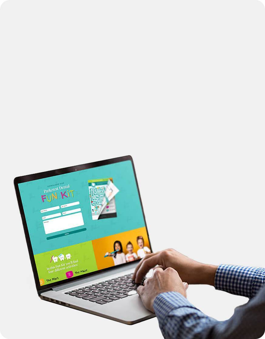A person in a blue checkered shirt types on a laptop displaying a colorful website labeled Fun Kit with login fields and images of children on the screen. The background is mostly white and empty.